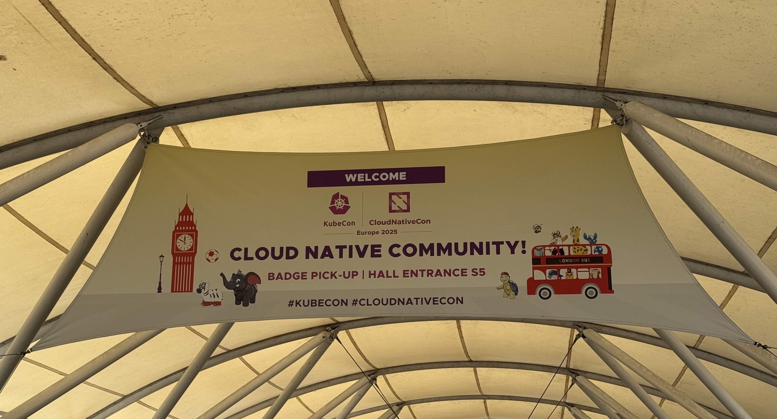 A KubeCon welcome banner mounted in the rafters of a tent-like entry 
