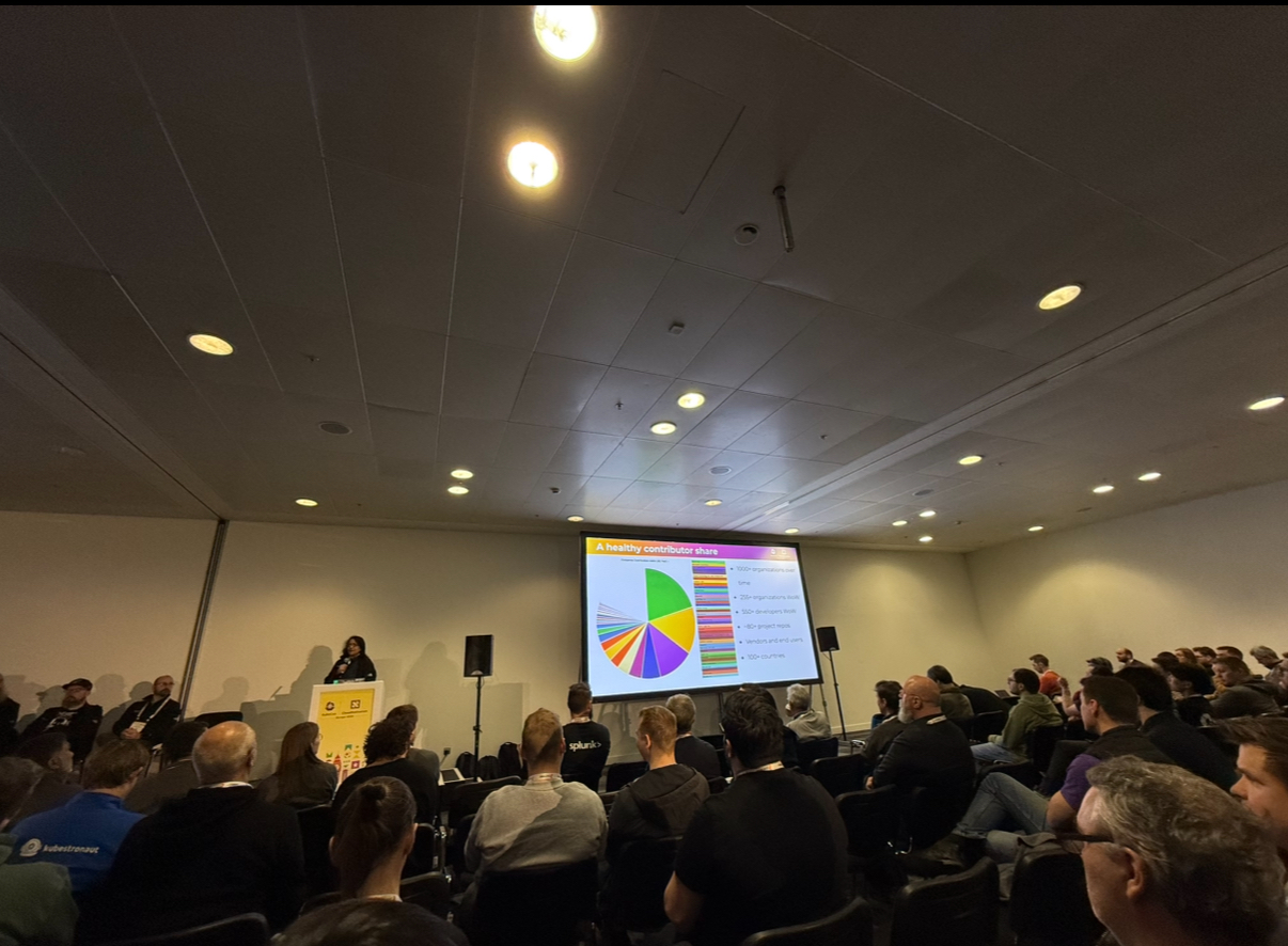 Photo of a conference room filled with people watching a speaker present OTel project updates slides displaying a pie chart. 