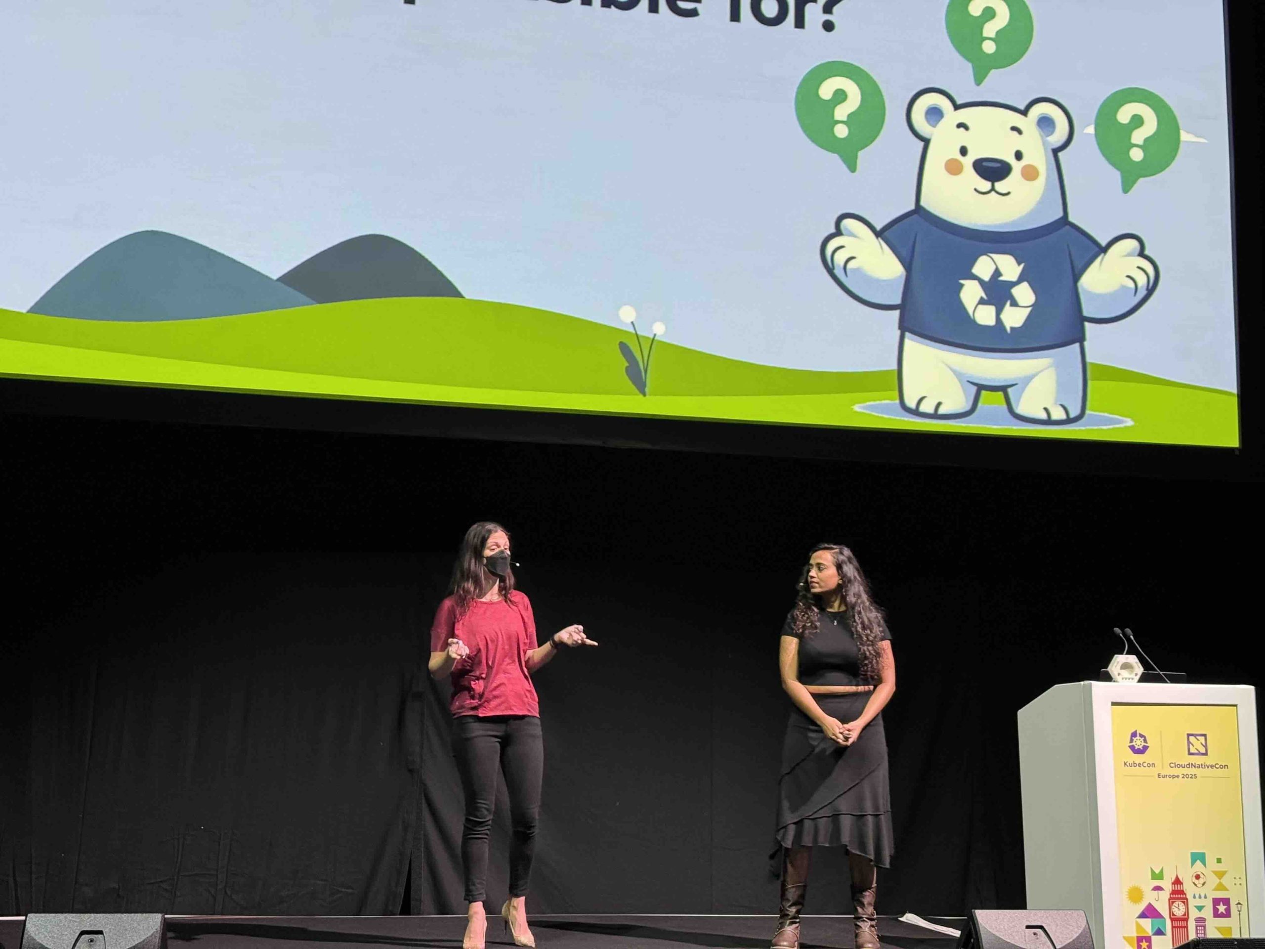 Nancy Chauhan and Adriana Villela on stage presenting a slide with a white bear wearing a recycling shirt at KubeCon EU 2025